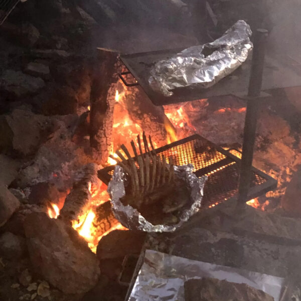 Meat wrapped in foil and a rib rack roasting over an open fire pit on metal grill racks during an Austere Cooking class at The Survival University.