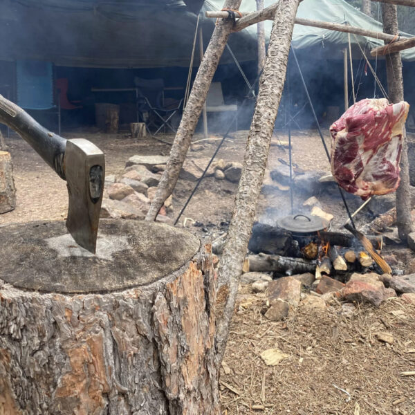 Axe embedded in a tree stump next to a campfire setup with a large cut of meat hanging from a wooden tripod and a lidded pot simmering over the flames during The Survival University’s Austere Cooking course.