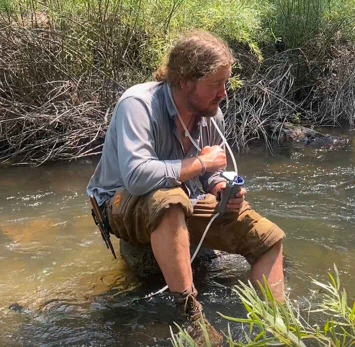 A man kneels on a rock in the middle of a shallow stream, using a portable water filter to safely drink from the flowing water. Dressed in outdoor gear with rolled-up sleeves and cargo shorts, he appears focused and calm, surrounded by lush greenery and rugged brush. This moment captures a practical skill in backcountry hydration during wilderness training.