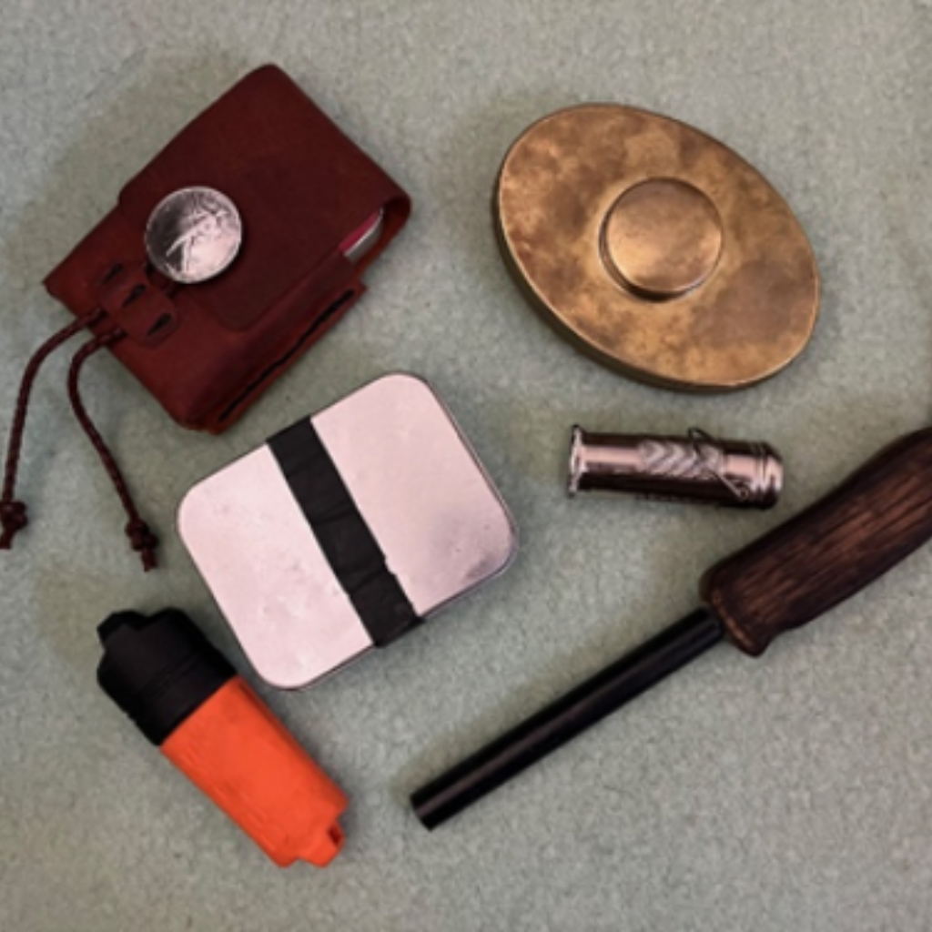 Assorted fire-starting tools including a ferro rod with wooden handle, waterproof match case, char tin, flint striker, brass tinder box, and leather pouch with a silver button