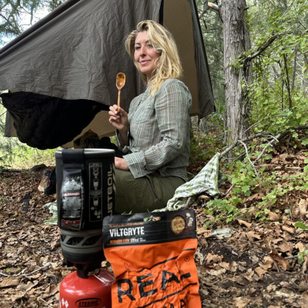 Jess sits on the forest floor beside a hammock shelter, holding a spoon and smiling at the camera. In the foreground are a Jetboil stove and a package of REAL Turmat freeze-dried meal.