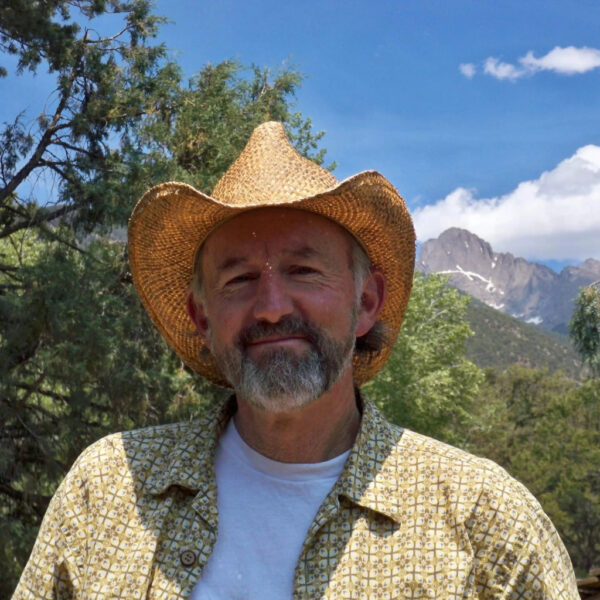 Cattail Bob stands outdoors with a calm smile, wearing a straw hat and patterned shirt, with Colorado mountain peaks and pine trees in the background under a bright blue sky.