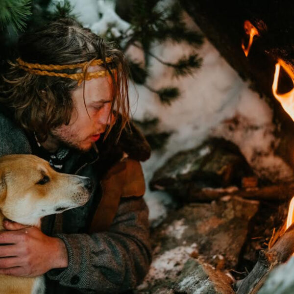 Ryan Maguire kneeling beside a fire in the snow, holding a dog close for warmth during a cold weather survival course.