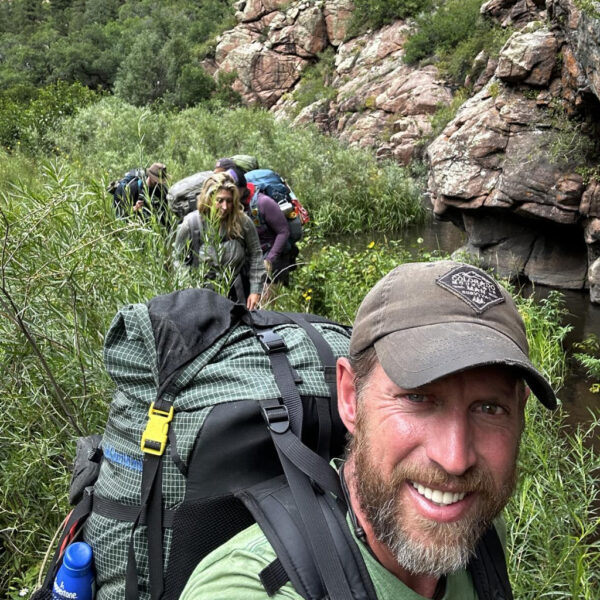 Jason takes a survival selfie while leading a group of hikers through lush greenery along a narrow trail beside a rocky mountain stream. All are wearing large backpacks and outdoor gear.