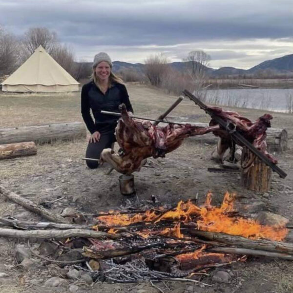 A woman, Bartle, kneels next to a fire pit in a grassy field with large animal quarters roasting on a wooden spit setup. A canvas tent stands in the distance near a lake, with a mountain range and cloudy sky in the background.
