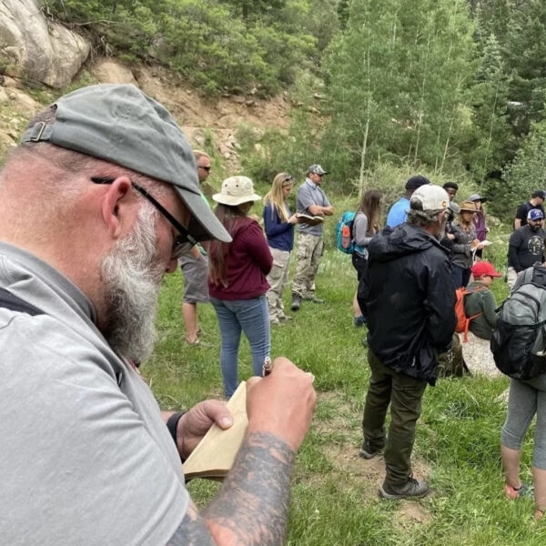 A tattooed man with a gray beard writes in a field notebook while a group of people gather in a lush mountain clearing for an outdoor class or nature walk.