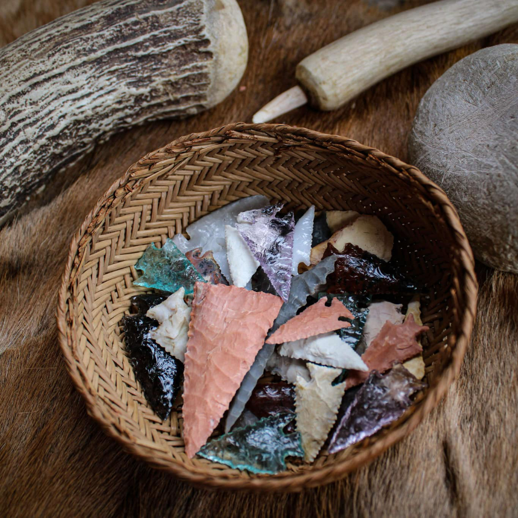 A woven basket filled with colorful hand-knapped stone arrowheads surrounded by traditional flint knapping