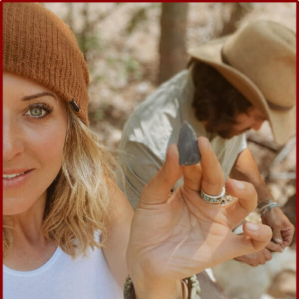 A woman in a white tank top and brown beanie holds up a small arrowhead toward the camera, smiling. A man in the background, wearing a tan hat, is focused on crafting or flint knapping.