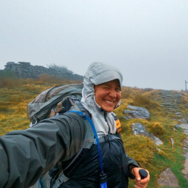 Jacks in a rain jacket winks and smiles while standing on a misty, stone-lined trail surrounded by wet grass and fog-covered hills.