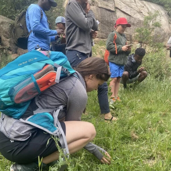 A group of participants kneels and crouches in a grassy meadow, closely examining wild plants during a foraging class. One woman in the foreground with a teal backpack inspects the ground while others observe and take notes.
