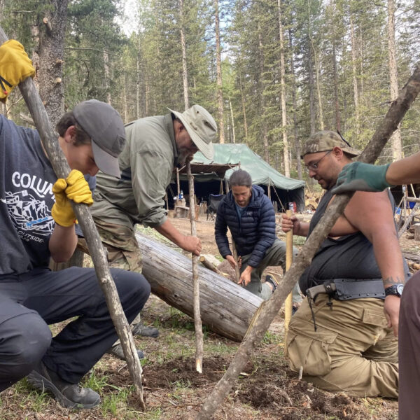 Group of students working together to lash poles and secure a log during a wilderness survival course at The Survival University, surrounded by forest shelters and tents