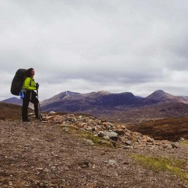 A solo hiker stands confidently at a rocky trail overlook, wearing a neon green jacket and carrying a large backpack. Dark hills and misty mountain peaks stretch across the horizon under a cloudy sky.