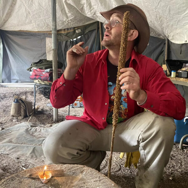 Hutch demonstrating how to use a dried mullein stalk as a primitive torch inside a canvas camp shelter during a survival course at The Survival University, with a small flame burning on a log stump in front of him.