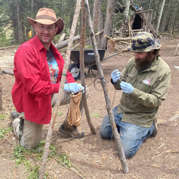 Hutch kneeling in the forest alongside another participant, demonstrating how to construct a tripod structure using cordage during a wilderness survival course at The Survival University.