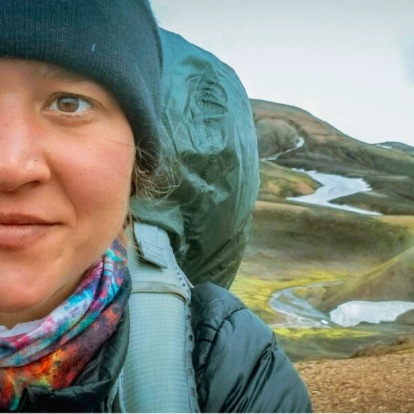 A close-up of a smiling hiker in a beanie and colorful neck gaiter, carrying a large backpack. Behind her are sweeping highland hills with winding snow patches and glacial ponds under a dramatic sky.