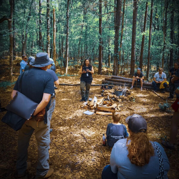 Jacks Genega stands at the center of a wooded clearing, teaching a group seated and standing around a fire pit during a wilderness class in the forest.