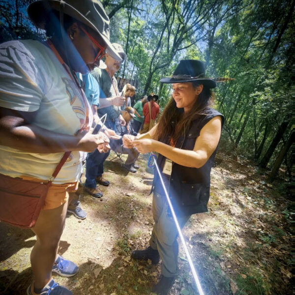 Jacks Genega smiles while teaching a student primitive skills during a hands-on wilderness class in a shaded forest clearing, with several participants engaged in learning behind them.