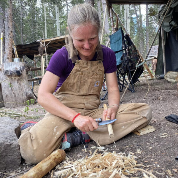 Participant sitting on the ground in a forest camp, smiling while carving fuzz sticks with a fixed-blade knife during a wilderness survival course at The Survival University.