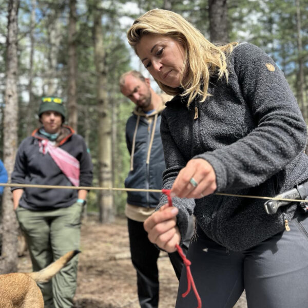 Jess demonstrates knot tying using red paracord on a line stretched between two trees, with several people and a dog watching in a forested area.