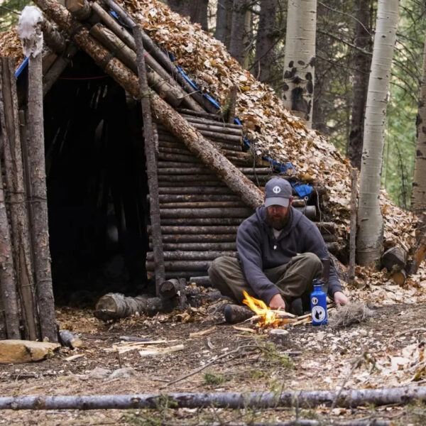 A man stands beside a partially enclosed primitive log shelter built from pine poles and forest debris in a dense pine forest. He is pointing toward the structure, which includes a raised bed area and a fire pit lined with stones.