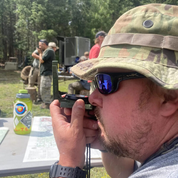 Man in camo hat uses lensatic compass during a navigation class, surrounded by others in an outdoor setting.