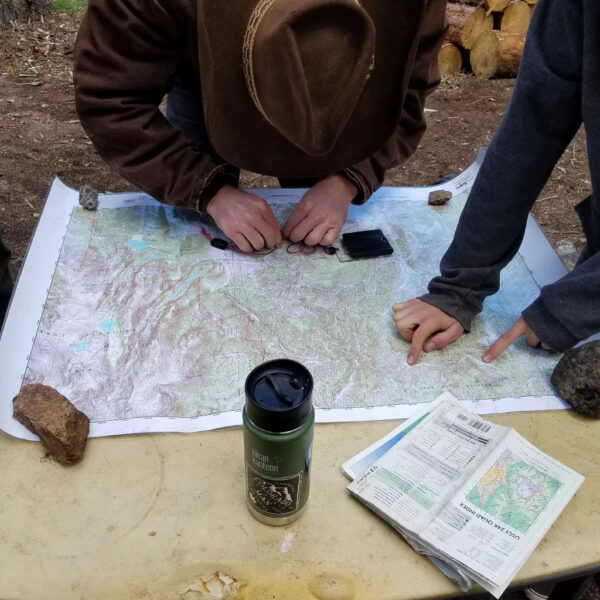 Two people mark a topographic map on a table using a compass, with navigation tools nearby.