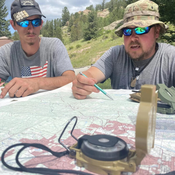 Two men in sunglasses study a topographic map using compasses and a pen at an outdoor navigation class.