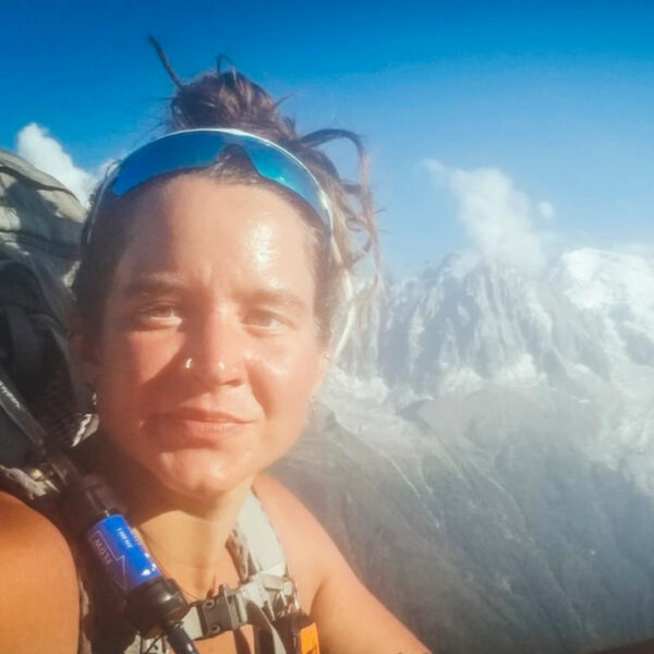Jacks with a hiking backpack and sunglasses poses for a selfie with a backdrop of snow-covered mountains under a clear blue sky.