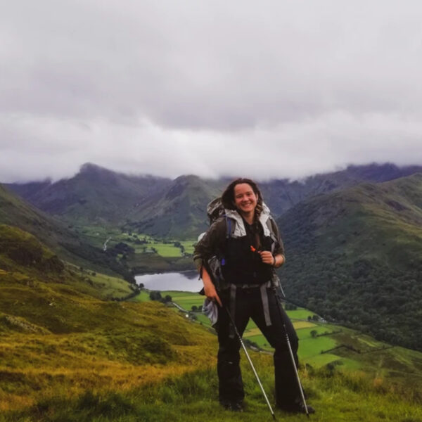 A smiling backpacker stands with trekking poles on a grassy mountain slope, overlooking a lush green valley with a reflective lake and misty peaks in the background.