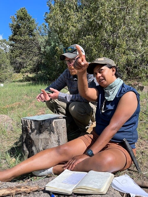 Matt Boger, land navigation instructor at The Survival University, sits with a student during an outdoor lesson. The student gestures toward a distant point while discussing navigation. They are seated in a forest clearing with notebooks, a protractor, and a map visible.