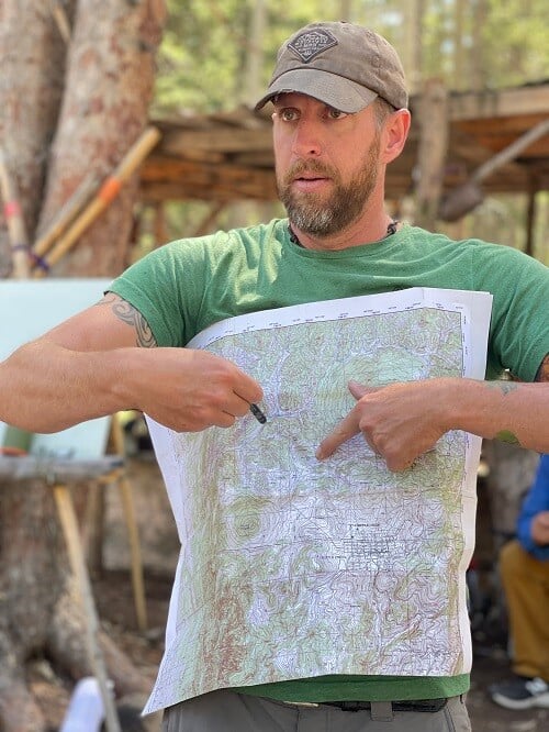 Jason Marsteiner holds up a topographic map during an outdoor class, pointing to elevation and contour lines as he explains map reading. Behind him, trees and wooden structures set the scene in a forest classroom environment.