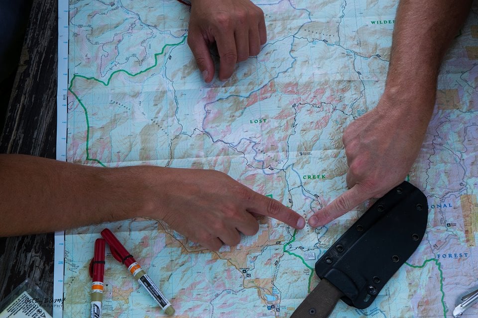 Two hands point to the same location on a topographic map during a land navigation exercise. The map shows mountainous terrain with labeled areas like 'Lost Creek.' Red markers and a sheathed knife rest nearby on the table.