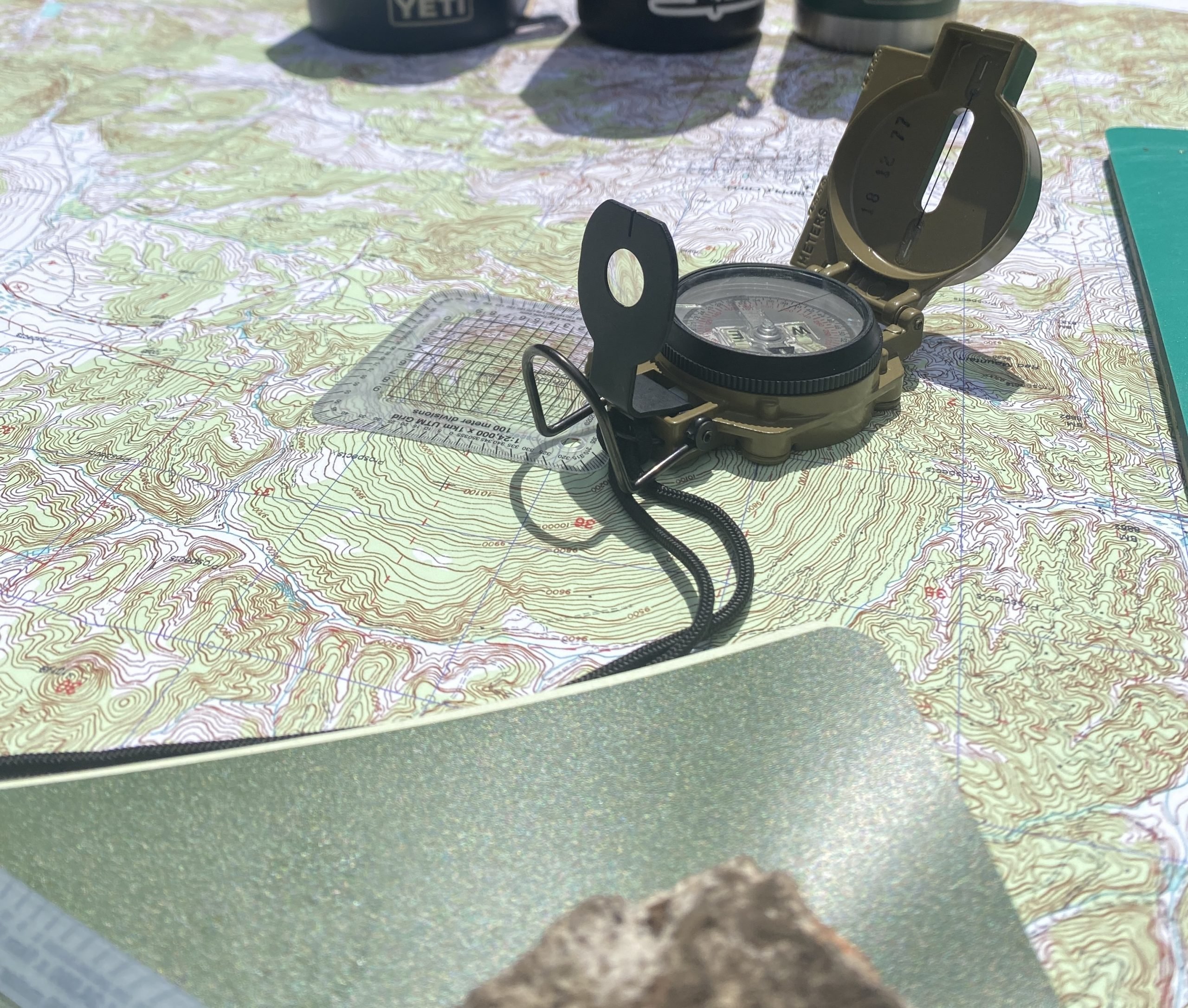 A military-style lensatic compass rests on a topographic map under bright sunlight, surrounded by navigation tools including a protractor and notebooks. Contour lines and elevation markings are clearly visible on the map.
