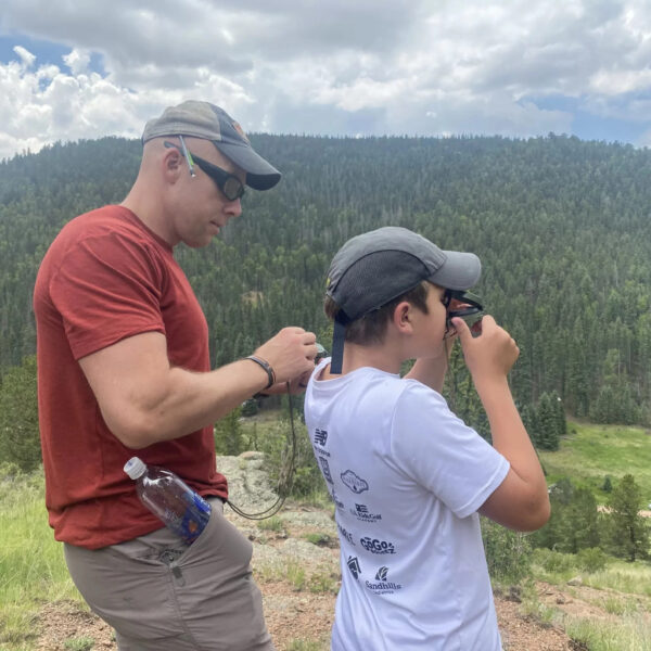 An instructor teaches a young student how to use a compass while overlooking a forested mountain landscape.