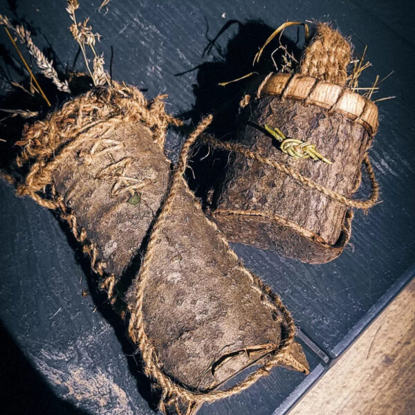 Two handmade bark containers wrapped in natural cordage, resting on a dark wooden surface. The containers are filled with dried grasses and herbs, showcasing primitive crafting techniques.