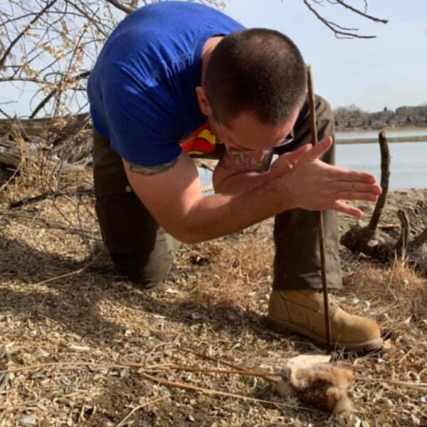 Hutch using the hand drill method to start a primitive fire near a riverbank during a wilderness survival course, pressing the spindle between his palms to create friction on a fire board.