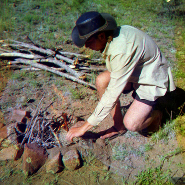 Man in hat kneeling to build a primitive fire with kindling and a ring of rocks in a grassy clearing.