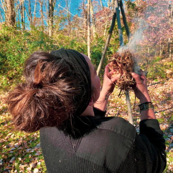 Jacks gently blows on a smoking tinder bundle to ignite a fire, surrounded by a sunlit forest covered in fallen leaves.