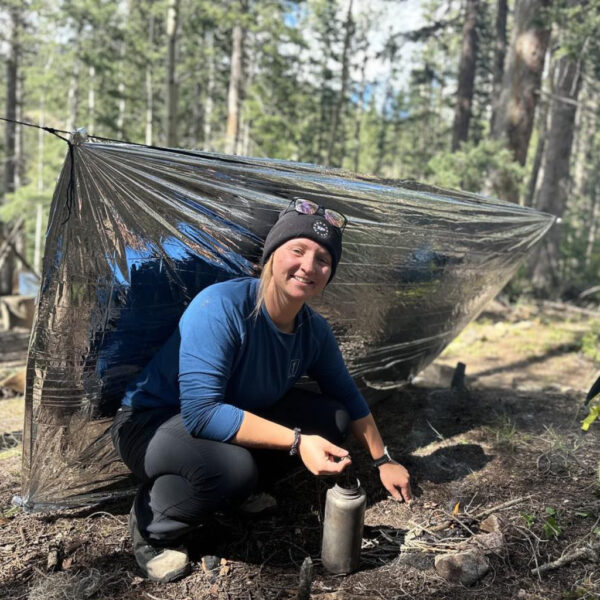 A woman crouches next to a small campfire, smiling, in front of a reflective emergency tarp shelter in a forest clearing. A backpack sits nearby.