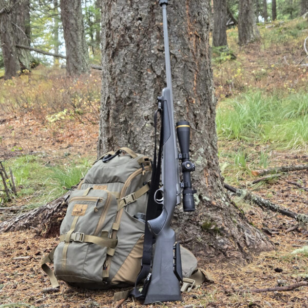 Scoped bolt-action rifle leaning on a backpack at the base of a tree in the forest.