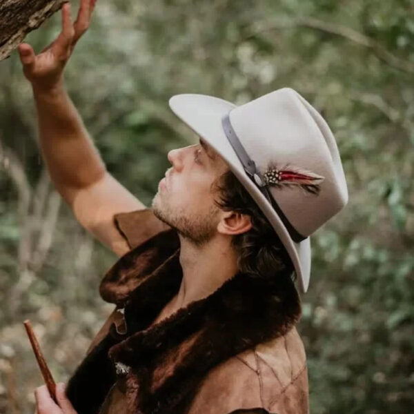 Ryan Maguire wearing a feathered hat and buckskin-style clothing, observing a tree closely while holding a wooden stick in a forest setting during a wilderness skills course.