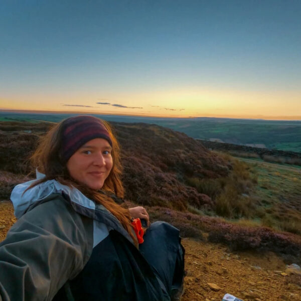 Jacks sits on a grassy ridge during golden hour, wearing a headband and windbreaker, with rolling hills and farmland stretching to the horizon under a glowing sunset sky.