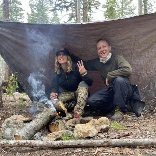 Two women sit under a tarp shelter in the forest, smiling near a smoky campfire made with large logs and rocks. One is flashing a peace sign while the other adjusts her hat.