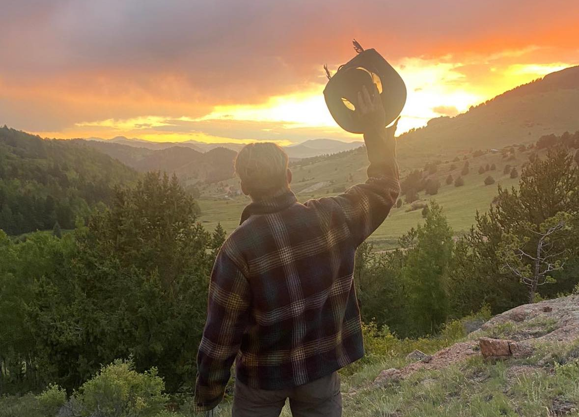 Person standing on a grassy hillside at sunset, facing away, holding a hat in the air and a water bottle in the other hand, with a dramatic sky glowing orange and purple over a mountainous landscape.