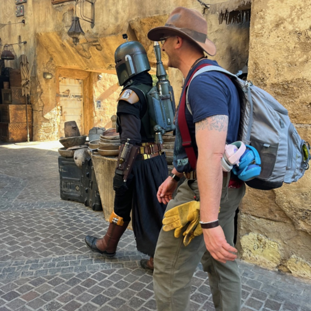 A man wearing a cowboy hat and backpack walks beside a Mandalorian cosplayer in a rustic Star Wars-themed marketplace