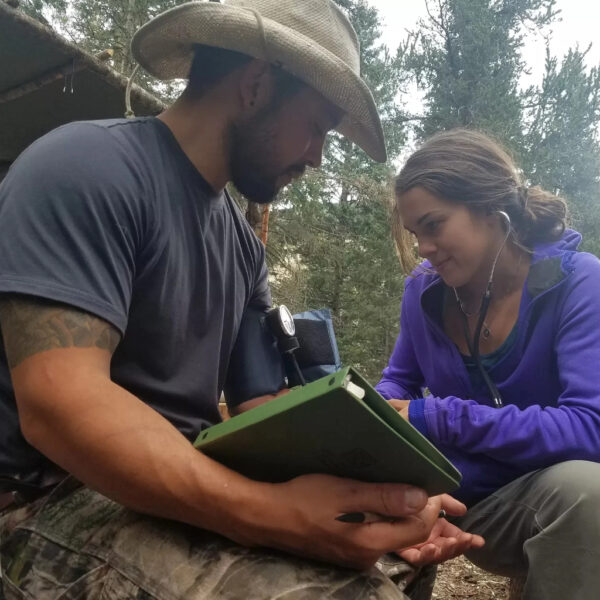 A woman in a purple hoodie uses a stethoscope to take a man's blood pressure during a wilderness first aid training session outdoors. The man is wearing a hat and holding a green notebook, surrounded by forest scenery.