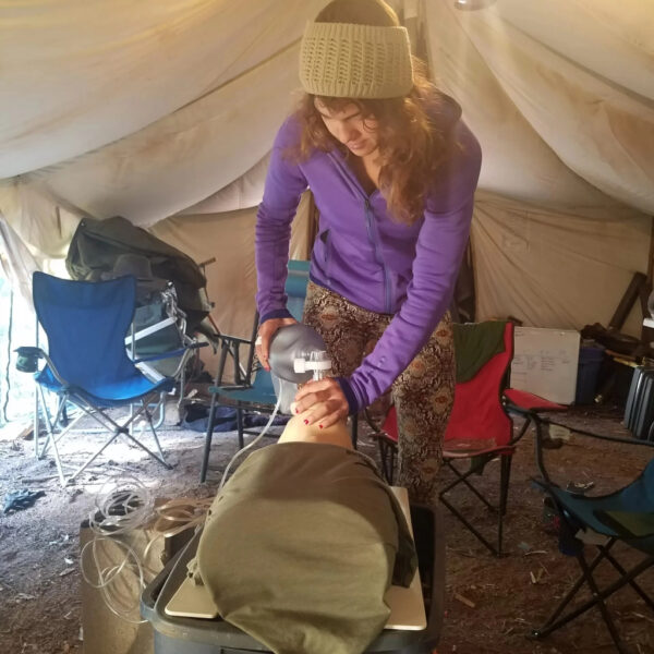 A woman in a purple hoodie and knit headband practices using a bag valve mask on a CPR training dummy inside a canvas tent. She is surrounded by camping chairs and medical training equipment, indicating a hands-on wilderness first aid session.