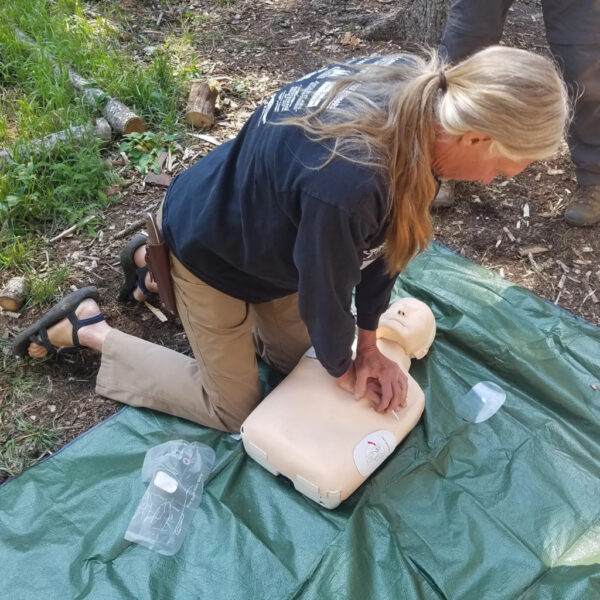 An instructor with long blond hair demonstrates CPR chest compressions on a training dummy during a Wilderness First Aid course. The session takes place outdoors on a green tarp surrounded by forest