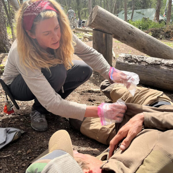 A woman wearing a red bandana and gloves provides first aid to a person lying on the ground during a Wilderness First Aid training in a forested area. She appears focused as she tends to the person's arm injury using a plastic bag and improvised materials.