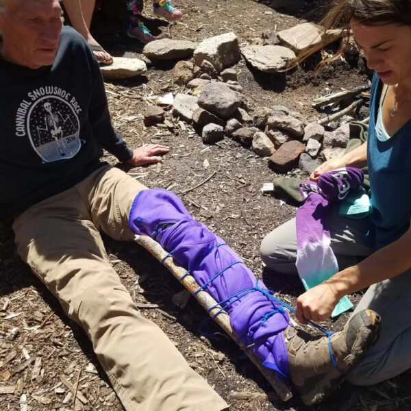 A woman in outdoor gear demonstrates how to apply a leg splint using a stick, purple fabric, and cordage on a seated man during a wilderness first aid class. They are seated on the forest floor near a fire pit.